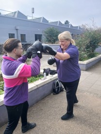Photograph of two people outdoors engaged in boxing training, with one person wearing black boxing gloves and the other holding focus mitts. Both individuals are smiling, dressed in casual clothing with purple tones, and standing near a low concrete wall with greenery and a building in the background.